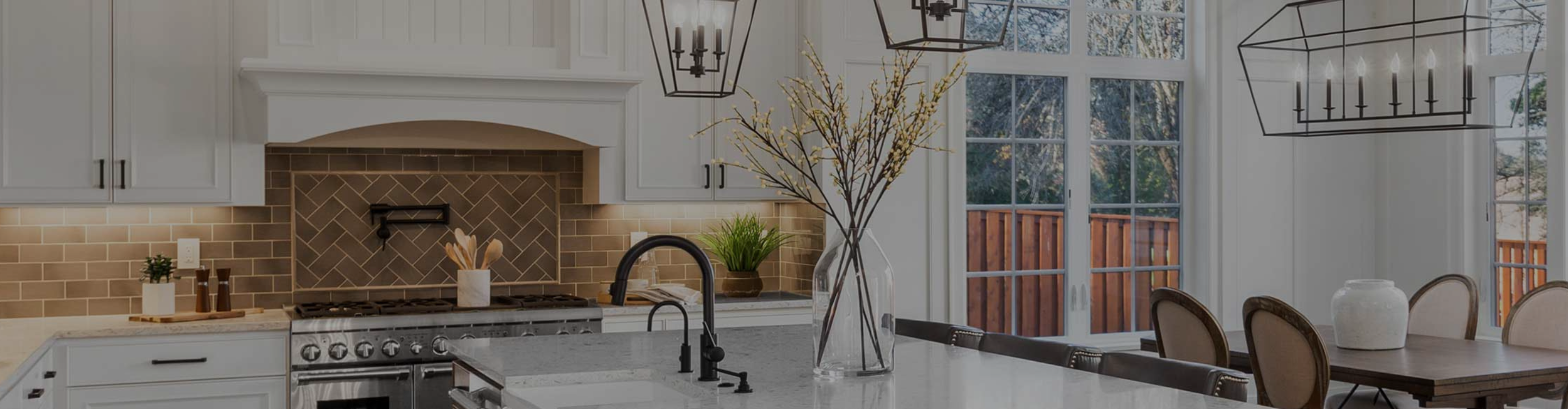 Modern kitchen with light cabinets, herringbone backsplash, stainless appliances, large island, black faucet, and warm lighting. Decorated with plants and a glass vase with blossoms.